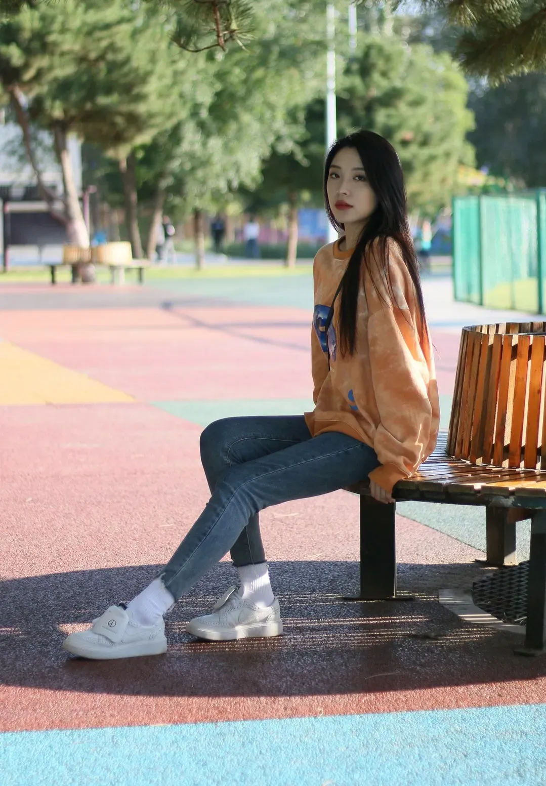 出镜人物身穿橙色套头卫衣与牛仔裤坐在室外弧形木长椅上