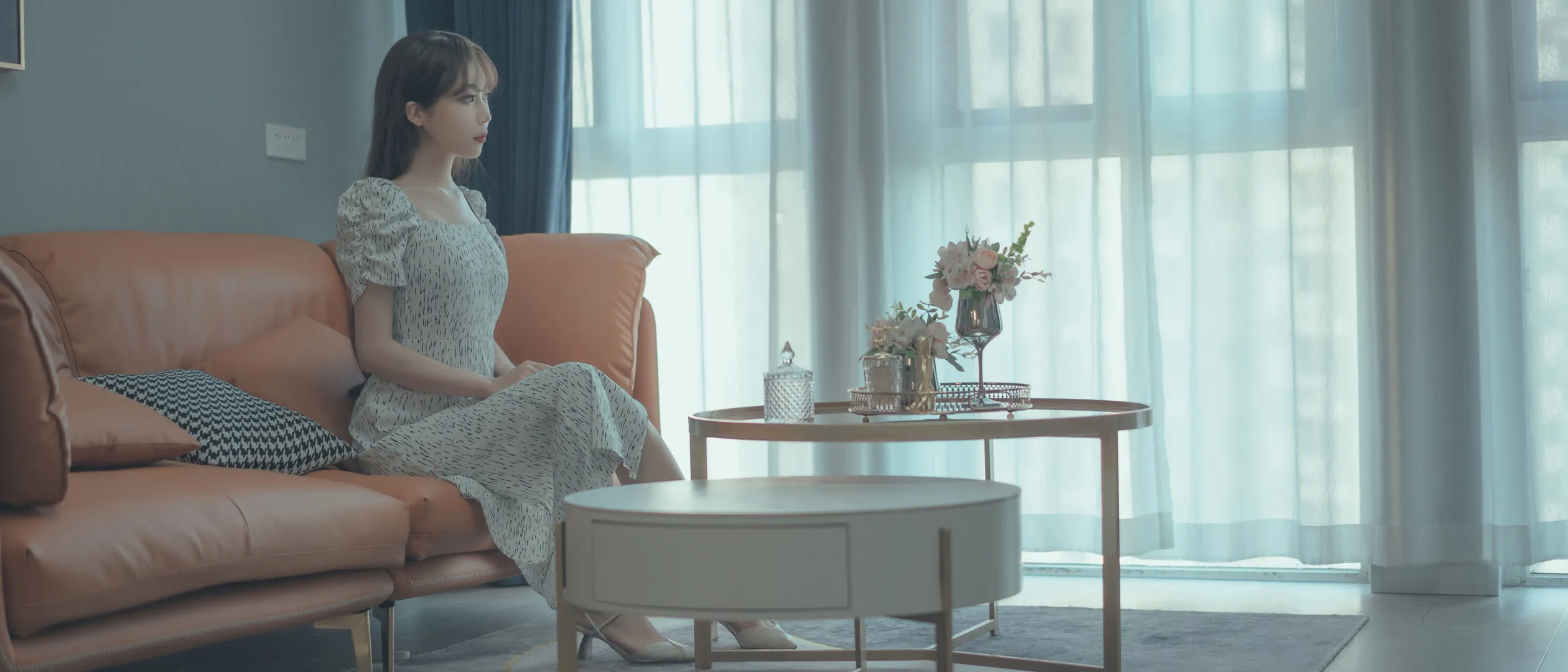 杏仁曲奇女仆造型与游戏角色写真风格参考 人物穿浅色连衣裙坐在橙色沙发旁,窗帘与茶几形成冷调室内场景